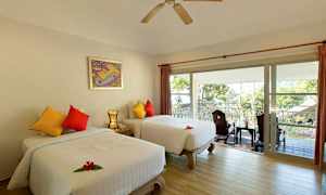 Et hotellrom med to separate senger, fargerike puter og en balkong med utsikt over trær og et tak utenfor. (AI-generert tekst)