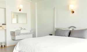 Et lyst hotellrom med en seng, grå puter og en vask med et speil. (AI-generert tekst)