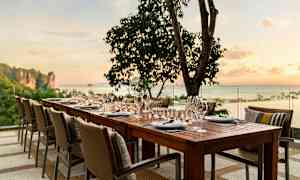 Et dekket bord med stoler står utendørs ved solnedgang med hav og trær i bakgrunnen. (AI-generert tekst)