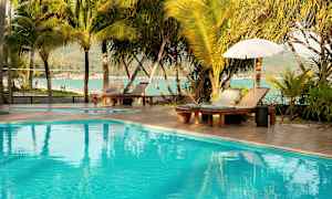 Et basseng med solsenger, parasoller og omkringliggende palmer ved sjøen. (AI-generert tekst)