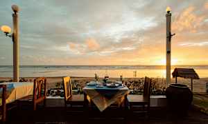 Utsikt over en strand ved solnedgang, med et dekket bord og stoler i forgrunnen. (AI-generert tekst)