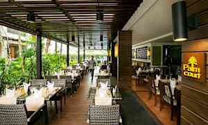 En restaurant med utendørs servering, møblert med kurvstoler og dekkede bord med duker og glass. (AI-generert tekst)