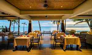 En restaurant ved sjøen med hvite duker på bordene og utsikt over palmer og stranden. (AI-generert tekst)