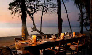 Langbord utendørs på en terrasse ved stranden, omgitt av trær og utsikt over havet i solnedgangen. Bordet er dekket med tallerkener, glass og lyslykter. (AI-generert tekst)