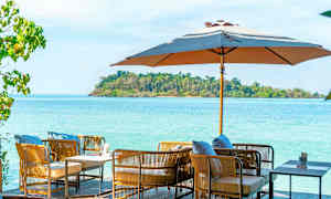 Utendørsmøbler med puter under en parasoll med hav og grønn øy i bakgrunnen. (AI-generert tekst)