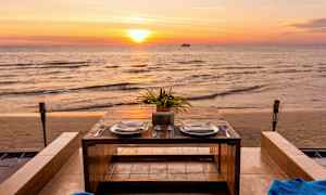 Et dekket bord med to tallerkener og bestikk står ved havet under en solnedgang. (AI-generert tekst)