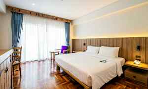 Et hotellrom med en dobbeltseng, mørke tregulv og et stort vindu med blå gardiner og hvit voile. (AI-generert tekst)