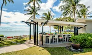 En utendørs bar ved en tropisk strand med palmer og hav i bakgrunnen. (AI-generert tekst)