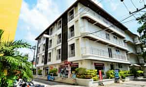 Bilde av en hvit bygning med flere etasjer, omgitt av grønne planter og motorsykler parkert ved siden av. (AI-generert tekst)