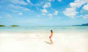 En kvinne går på en lys sandstrand med havet og en blå himmel i bakgrunnen. (AI-generert tekst)