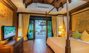 Et soverom med trekonstruksjon, som har utsikt mot en terrasse og havet utenfor. Rommet inneholder en seng med fargerike puter, et TV-skap og lamper. (AI-generert tekst)