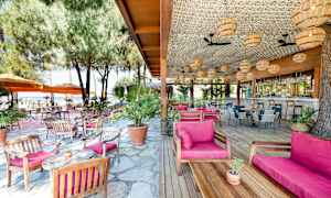 Utendørsrestaurant med trebord og stoler, rosa puter, omgitt av trær og parasoller. (AI-generert tekst)