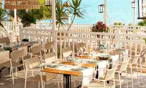 En utendørs restaurant med bord dekket med tallerkener og glass. Utsikt over havet i bakgrunnen. (AI-generert tekst)
