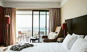 Et hotellrom med en stor seng, en lenestol og en balkong med utsikt mot havet. (AI-generert tekst)