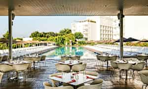 Bilde av en utendørsrestaurant med bord og stoler, ved et stort basseng omgitt av solsenger og parasoller, med grøntområder og bygninger i bakgrunnen. (AI-generert tekst)