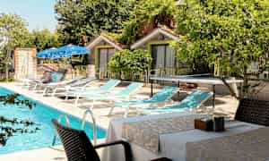 Et utendørs svømmebasseng ved siden av solsenger, bord og stoler plassert nær små bygninger. (AI-generert tekst)