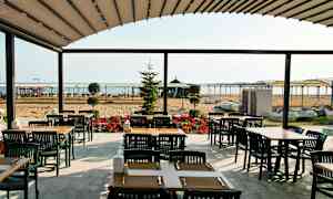 En utendørs servering med bord og stoler, i bakgrunnen en sandstrand og havet. (AI-generert tekst)