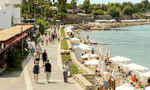 En gangsti ved havet med folk som spaserer og personer som slapper av på solsenger under parasoller. (AI-generert tekst)