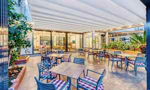 Bilde av en utendørsterrasse med bord og stoler, samt planter og et innglasset innendørsområde i bakgrunnen. (AI-generert tekst)