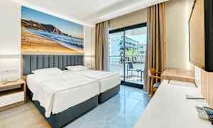 Hotellrom med to enkeltsenger, balkong i bakgrunnen og stort fotografi av en strand på veggen. (AI-generert tekst)