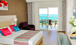 Hotellrom med dobbeltseng, fargerike puter og utsikt mot en balkong med havutsikt. (AI-generert tekst)