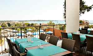 Utsikt fra en restaurant med dekkede bord og stoler, uteområde med parasoller og havet i bakgrunnen. (AI-generert tekst)