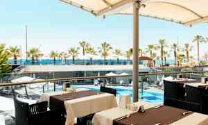 Utsikt fra en utendørs servering mot et bassengområde med parasoller og palmer samt havet i bakgrunnen. (AI-generert tekst)