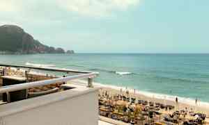 Et bilde av en strand med solsenger, parasoller og mennesker som svømmer i havet. I bakgrunnen vises en fjellklippe. (AI-generert tekst)