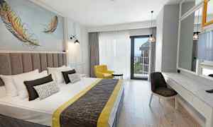 Et hotellrom med en dobbeltseng, gul lenestol, skrivebord og speil, samt balkong med havutsikt. (AI-generert tekst)