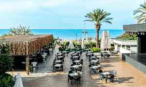 En utendørsrestaurant med bord og stoler, plassert nær havet. (AI-generert tekst)