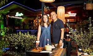 To personer ved et dekket bord med en kake og tente lys på en restaurant med grøntområde og lamper. (AI-generert tekst)