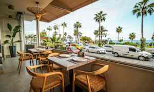 Bildet viser en restaurant med utsikt mot en gate med parkerte og forbipasserende biler samt palmer i bakgrunnen. (AI-generert tekst)