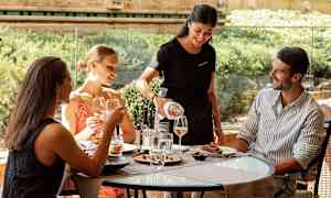 En servitør serverer drikke til gjester som sitter ved et bord på en uterestaurant. (AI-generert tekst)