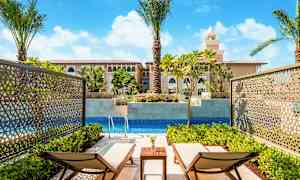 To solsenger ved et privat basseng, omgitt av grønne planter og med utsikt mot en bygning med middelhavsinspirerte arkitekturelementer. (AI-generert tekst)