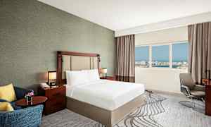 Et hotellrom med en dobbeltseng, nattbord, lenestol og vindu med utsikt over en by. (AI-generert tekst)