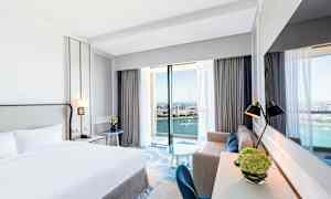 Hotellrom med dobbeltseng og havutsikt gjennom store vinduer. (AI-generert tekst)