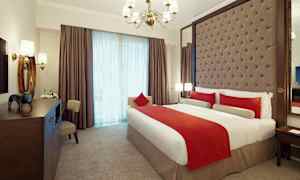 Et elegant innredet hotellrom med en stor seng kledd i hvitt og rødt sengetøy. Rommet har en polstret sengegavl, en lysekrone og store vinduer med gardiner. (AI-generert tekst)