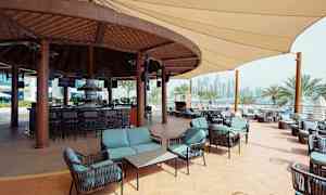 En utendørs bar med sittegrupper og tak i tre, omgitt av palmer og byens skyline i bakgrunnen. (AI-generert tekst)