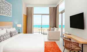 Et moderne hotellrom med en stor seng, en sofa, skrivebord og en TV. Utsikten fra rommet viser en strand og havet. (AI-generert tekst)