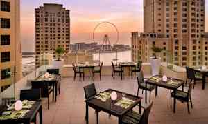 En terrasse med bord og stoler, omgitt av høye bygninger med utsikt over et pariserhjul og havet ved solnedgang. (AI-generert tekst)