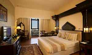 Et hotellrom med en dobbeltseng, skrivebord og sittegruppe. Vinduet har utsikt med dekorative detaljer. (AI-generert tekst)