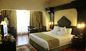 Et hotellrom med en stor seng, mørke tremøbler og vinduer dekket med elegante gardiner. (AI-generert tekst)