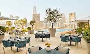 En utendørsterrasse med bord og stoler med utsikt over et basseng og byens silhuett i bakgrunnen. (AI-generert tekst)