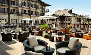 En utendørsterrasse med moderne sitteplasser, parasoller og planter, omgitt av bygninger. (AI-generert tekst)