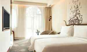 Et hotellrom med to senger dekket med hvite laken. Et sitteområde med et lite bord og vindu med tynne gardiner vises i bakgrunnen. (AI-generert tekst)