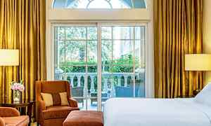 Et hotellrom med store vinduer som vender mot en terrasse med utsikt over grønt område. Rommet har en seng, lenestoler og gullfargede gardiner. (AI-generert tekst)