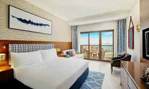 Et hotellrom med en dobbeltseng, sofa og balkong med utsikt mot havet. (AI-generert tekst)