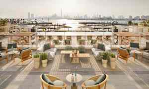 En utendørsterrasse med møbler og utsikt over en havn og byens silhuett i bakgrunnen. (AI-generert tekst)