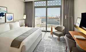 Et moderne innredet hotellrom med en stor dobbeltseng og vinduer som åpner opp til en balkong med utsikt over en bysilhuett og vann. (AI-generert tekst)