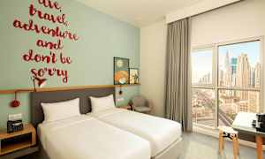 Et hotellrom med to enkeltsenger, en veggdekorasjon med et inspirerende sitat, og et vindu med utsikt over byens silhuett. (AI-generert tekst)